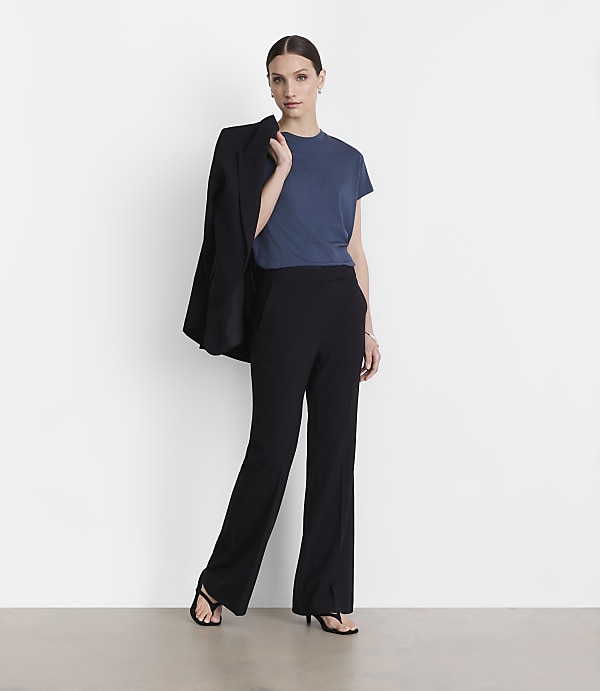 A woman in a blue t-shirt and black pants stands against a white background, with a black blazer draped over her shoulder.