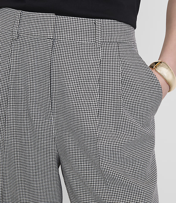 A person's hand, adorned with a gold bracelet, rests in the pocket of houndstooth patterned pants against a plain white background.