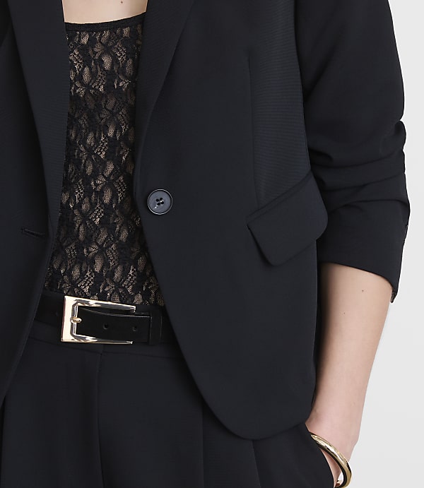 A woman in a black blazer and lace top, with a black belt and gold bracelet, stands against a plain white background.
