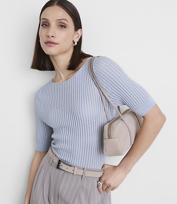 A woman in a light blue ribbed top and pinstripe pants poses with a small beige handbag against a plain white background.