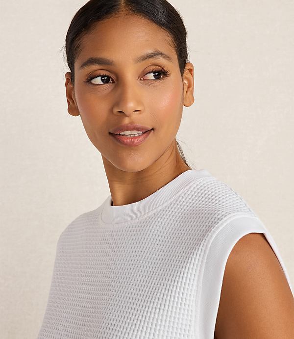 A woman with dark hair pulled back smiles softly, wearing a white waffle-knit top against a plain, light background.