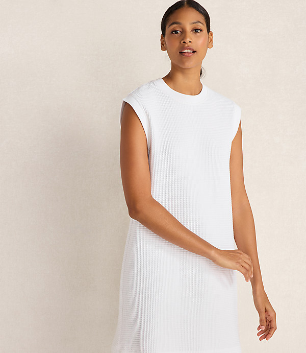 A woman in a white waffle-knit dress stands against a textured, light-colored wall.
