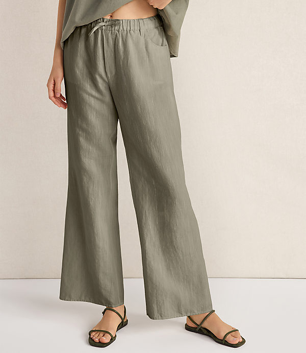 A person wearing loose-fitting, olive-green linen pants and sandals stands against a plain, textured background.