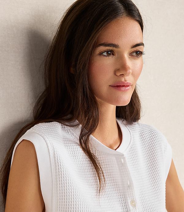 A young woman with long, dark hair wears a white waffle-knit top in the foreground, with a textured, light-colored wall in the background.