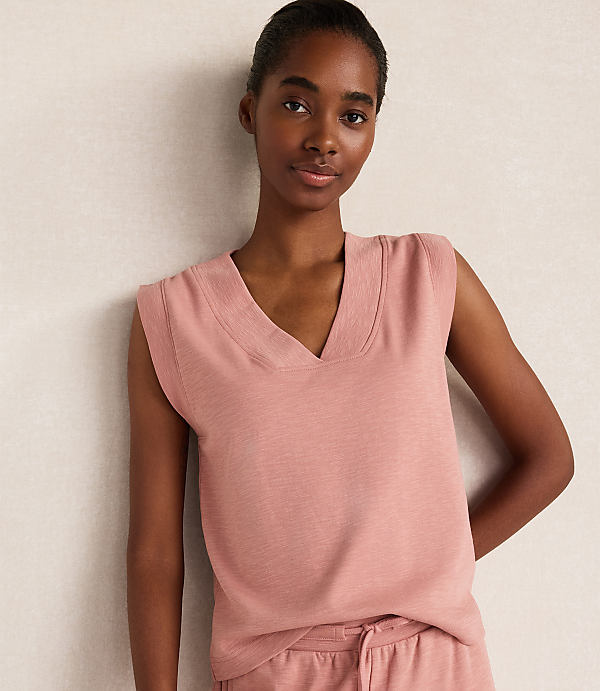 A young woman in a pink v-neck sweater vest stands against a textured, light-colored wall.