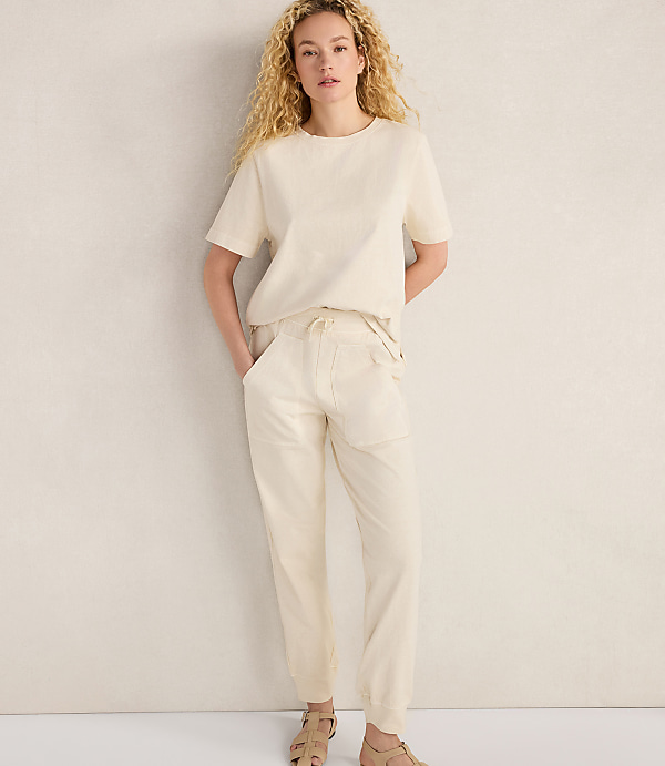 A woman in a cream-colored t-shirt and pants stands against a plain, light-colored wall.