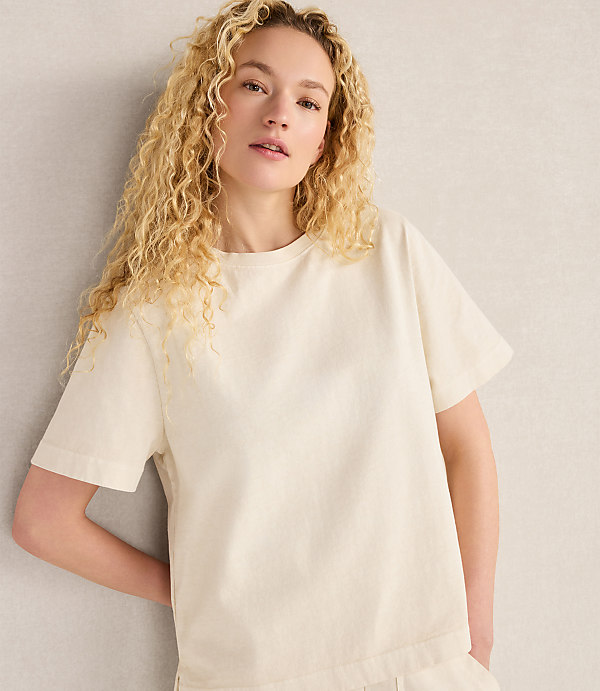 A young woman with curly blonde hair wears a cream-colored t-shirt, leaning against a neutral-toned wall.