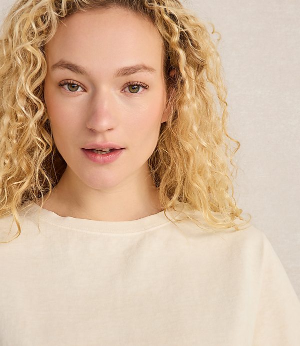 A young woman with curly blonde hair and fair skin looks forward against a textured, light-colored background.