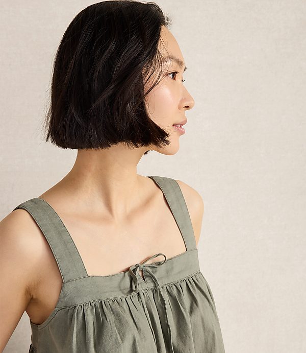 A woman with short dark hair wears a green sleeveless top with a tie at the neckline, set against a plain, light-colored background.