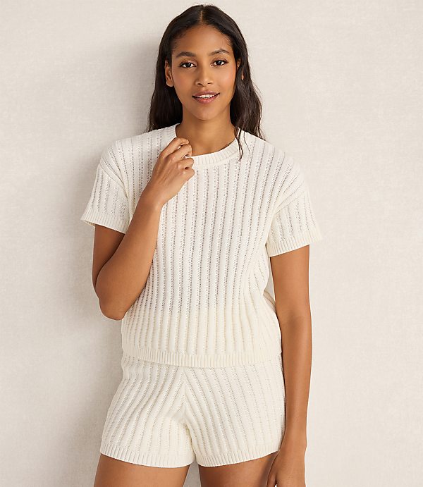 A woman in a cream-colored cable-knit top and shorts stands against a textured, light-colored background.