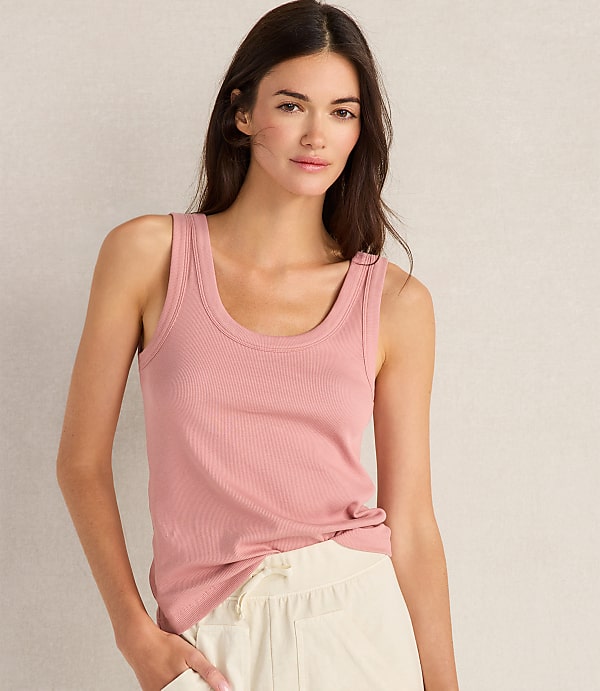 A woman in a pink ribbed tank top and cream-colored pants stands against a plain, light-colored background.