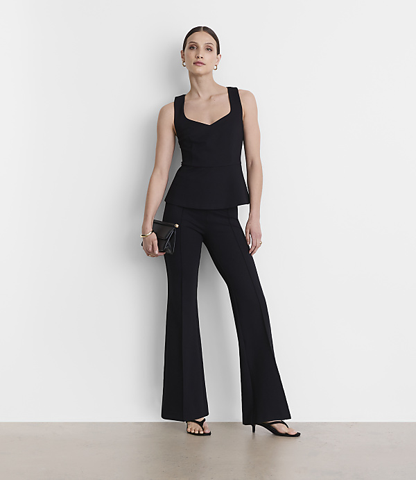 A woman in a black peplum top and flared pants stands against a plain white background, holding a black clutch.