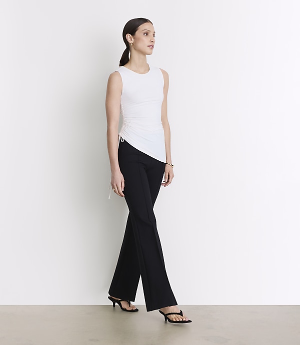 A woman in a white sleeveless top and black pants stands against a plain white background.