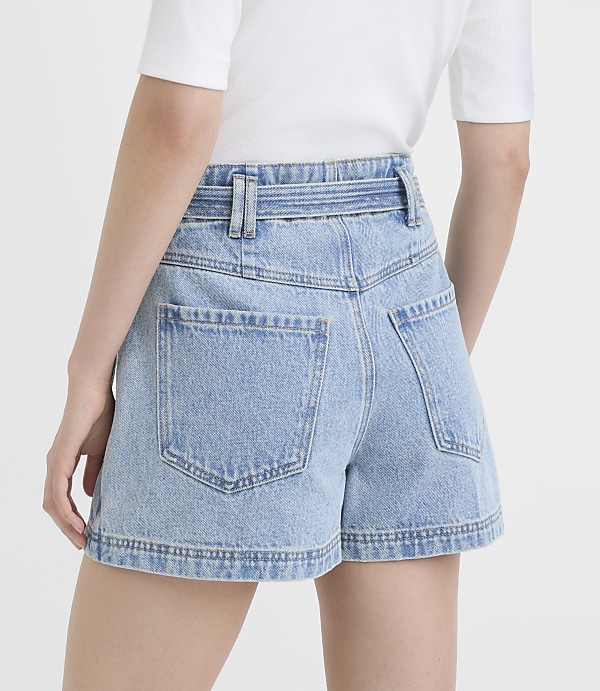 A person wearing light-wash denim shorts with a white belt stands against a plain white background.