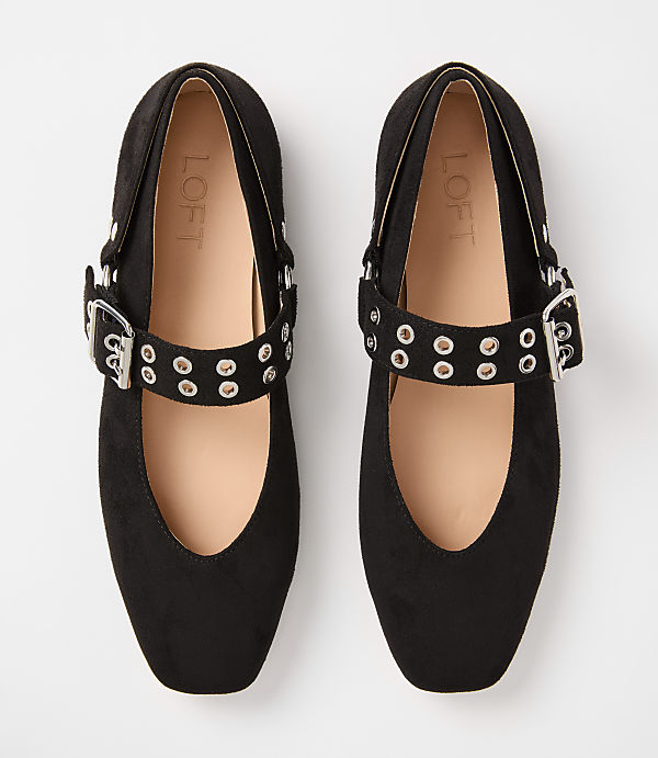 A pair of black suede Mary Jane flats with silver buckle straps and grommets rests on a plain white background.