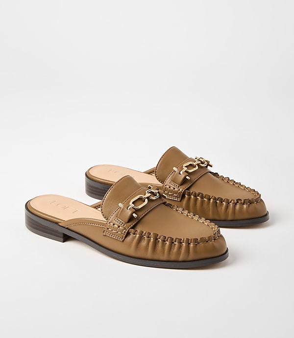 A pair of tan leather mules with gold chain accents rests on a plain white background.