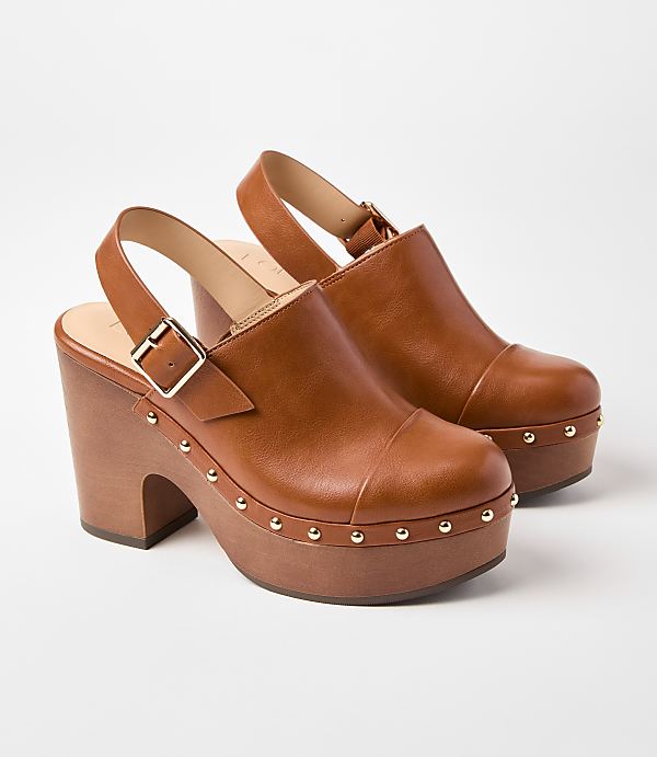 A pair of brown leather clogs with wooden platform soles and gold stud detailing are displayed against a plain white background.