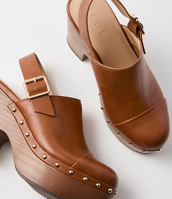 Two brown leather clogs with wooden heels and brass stud detailing are arranged on a plain white background.