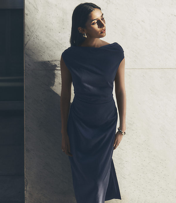A woman in a dark blue dress stands against a marbled wall.