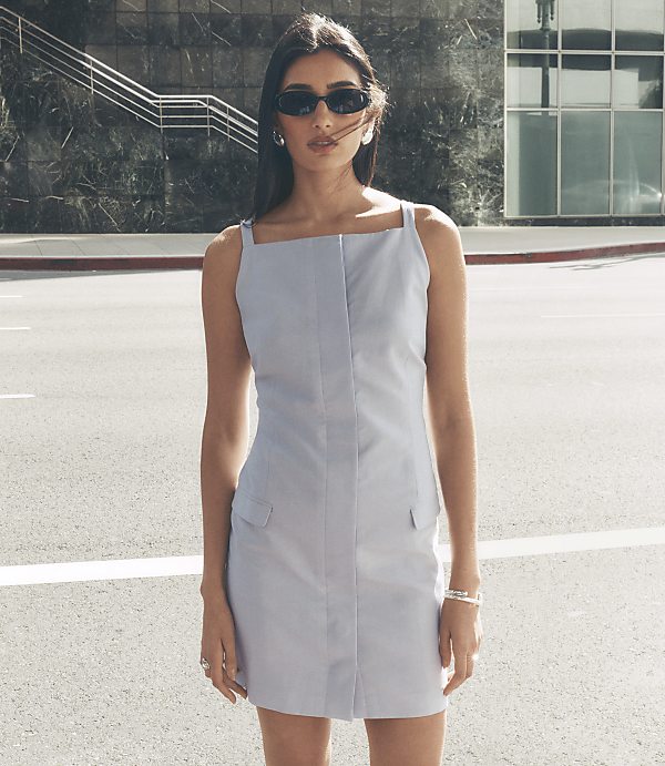A woman in a light blue dress and sunglasses stands on a street with a modern building in the background.