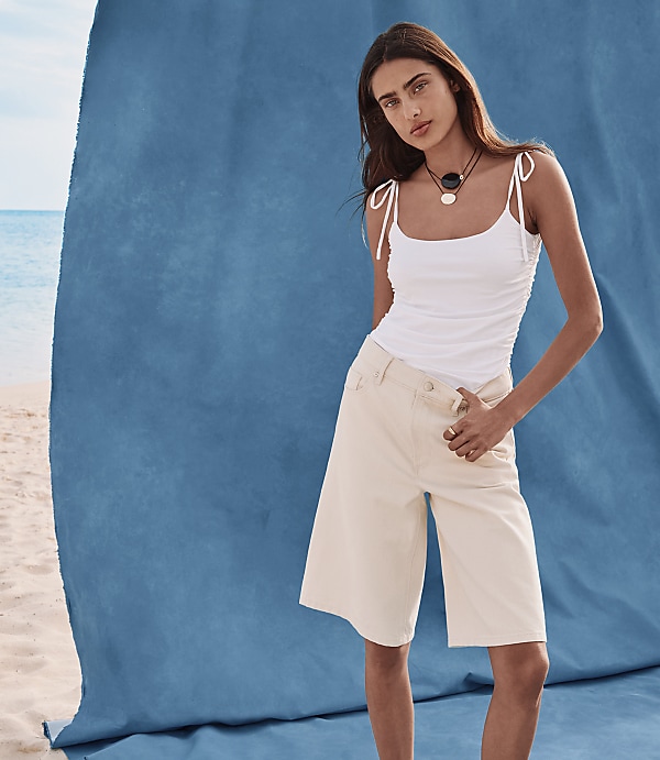 A woman in a white tank top and beige culottes stands on a sandy beach in front of a large blue backdrop.