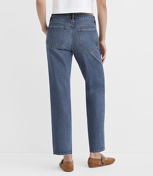 A person wearing blue jeans and brown loafers stands against a plain white background.