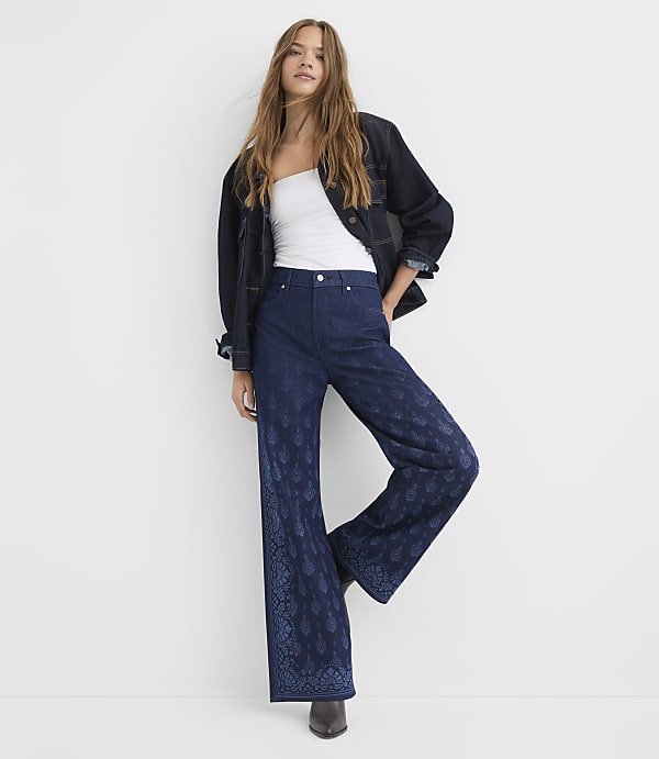 A woman in a white tube top, dark denim jacket, and patterned wide-leg jeans stands against a plain white background.