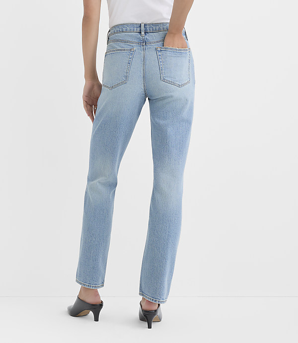 A person wearing light-wash denim jeans and black heeled mules stands against a plain white background.