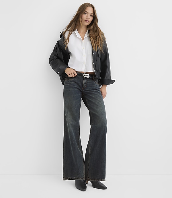 A woman in a white shirt, black jacket, and wide-leg jeans stands against a plain white background.
