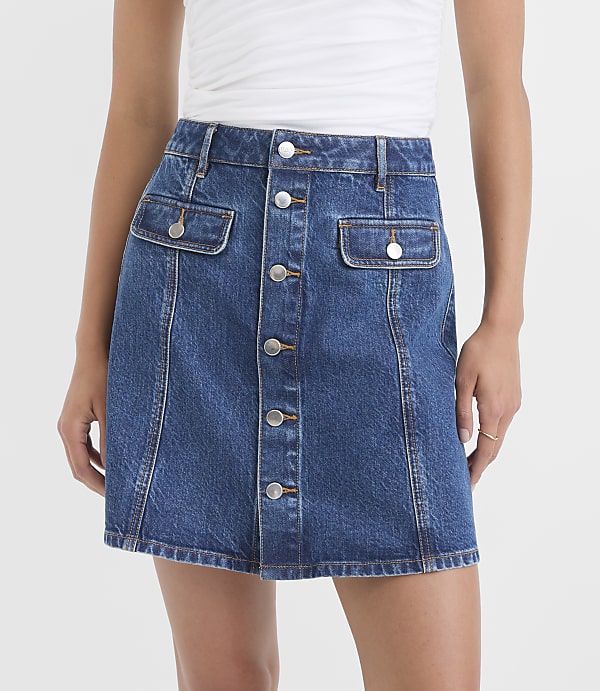 A woman in a white top wears a blue denim A-line skirt with a button-front closure and two chest pockets, set against a plain white background.