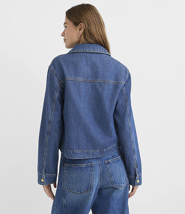 A woman in a denim jacket and jeans stands against a plain white background.