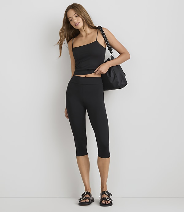 A woman in a black tank top and capri leggings stands against a plain white background, carrying a black tote bag.