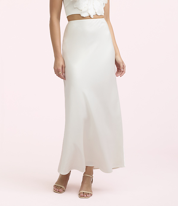 A woman in a white satin skirt and sandals stands against a soft pink background.