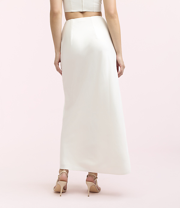 A woman in a white skirt and heels stands against a plain pink background.