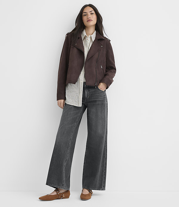 A woman in a brown jacket, striped shirt, and wide-leg jeans stands against a plain white background.