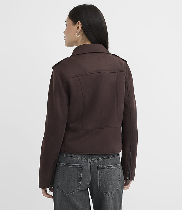 A woman in a brown jacket and jeans stands against a plain white background.
