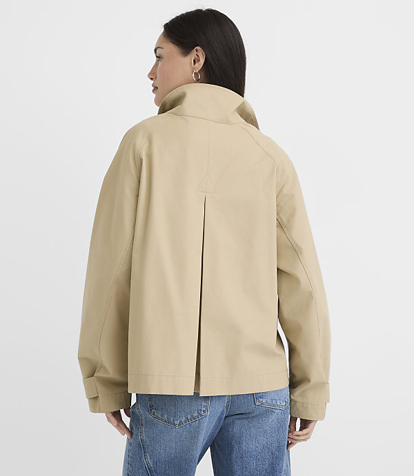 A woman in a beige jacket and blue jeans stands against a plain white background.