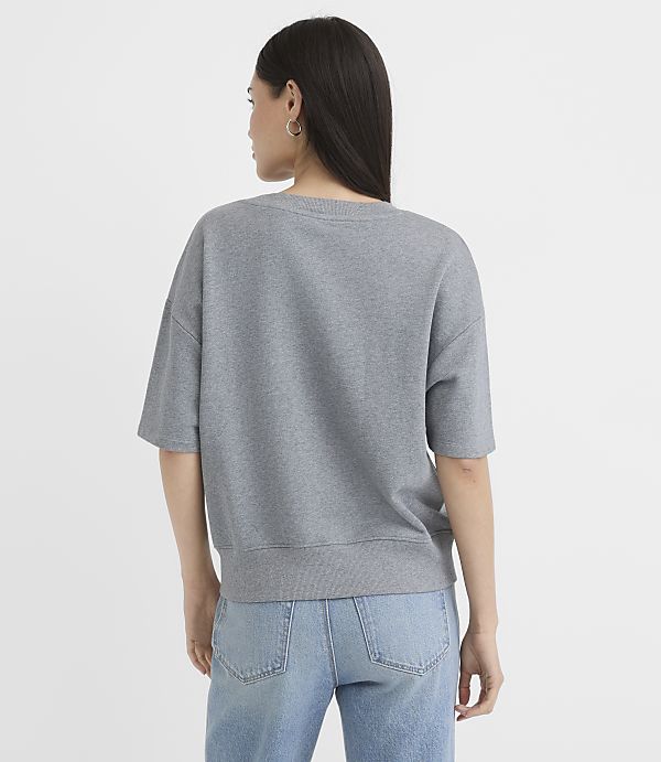 A woman in a grey sweatshirt and blue jeans stands against a plain white background.