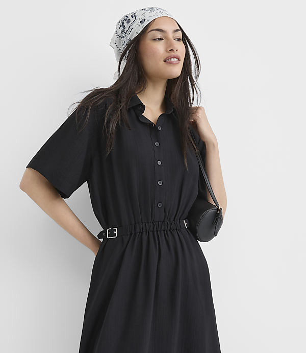 A woman in a black dress and bandana poses against a plain white background, with a small black purse slung over her shoulder.