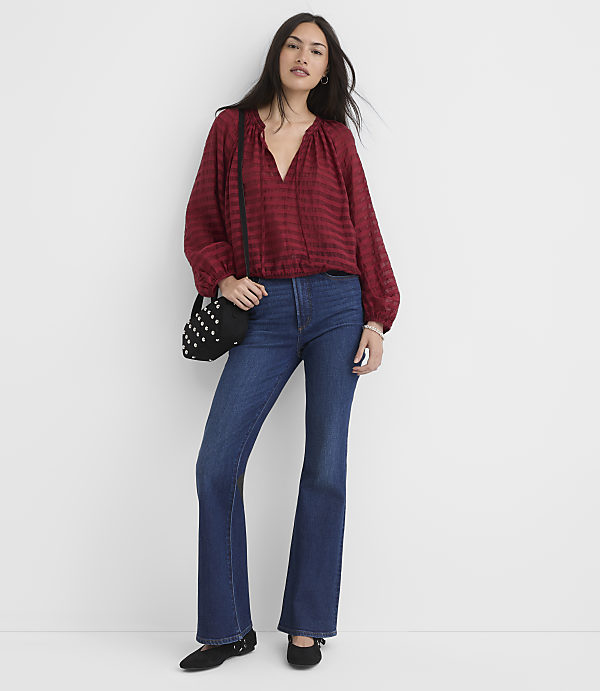 A woman in a red blouse and flared jeans stands against a plain white background, accessorized with a studded black bag and black flats.