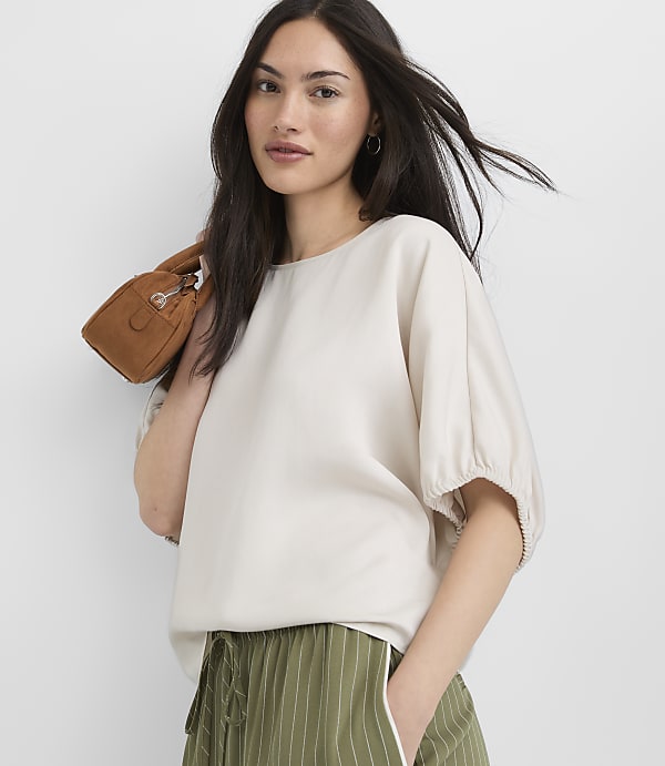 A woman in a cream top and green striped pants holds a small brown purse in front of a plain white background.
