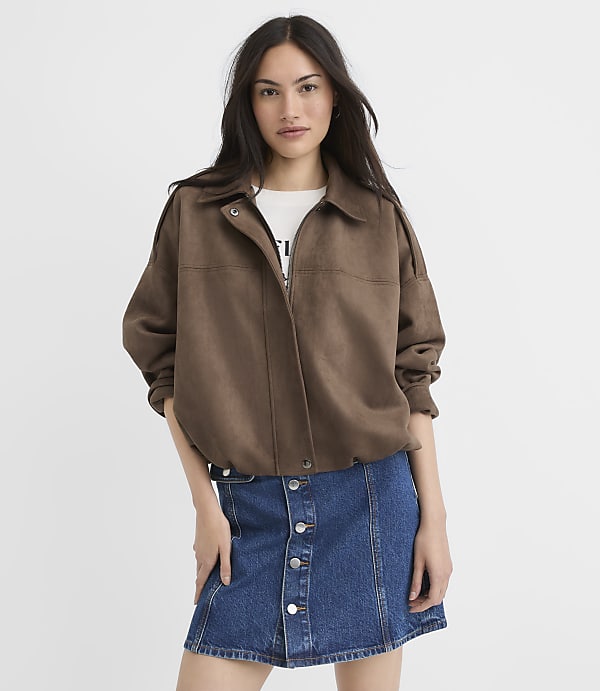 A woman in a brown jacket and denim skirt stands against a plain white background.