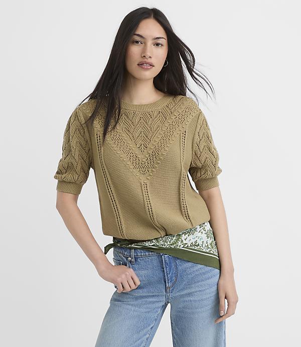 A woman in a textured olive green sweater and light wash jeans stands against a plain white background.