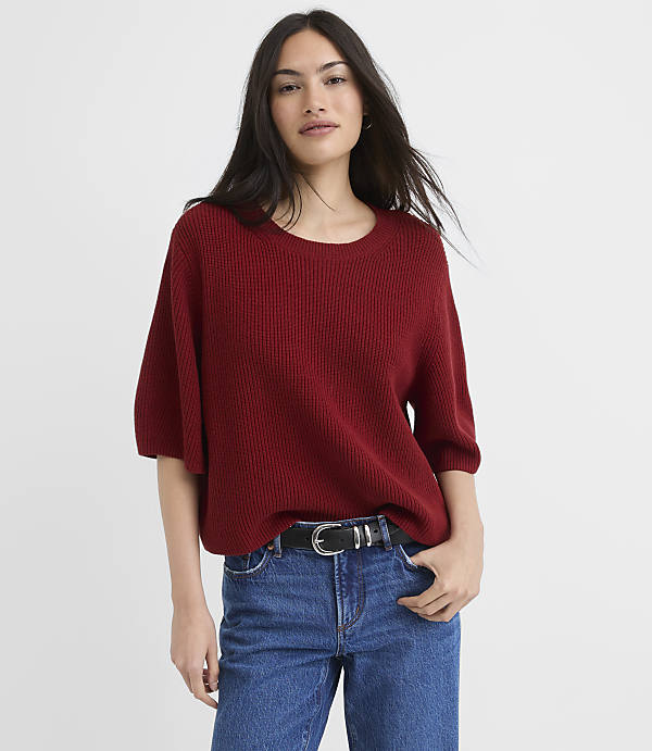 A woman in a red sweater and blue jeans stands against a plain white background.