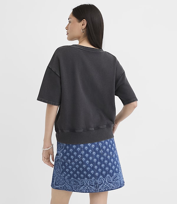 A woman in a dark gray t-shirt and a blue patterned skirt stands against a plain white background.