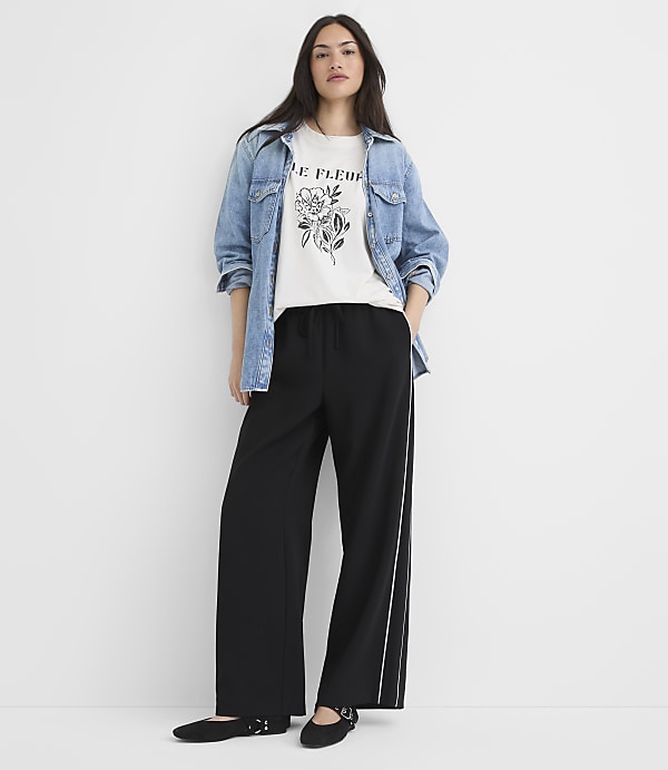 A woman in a graphic tee, denim jacket, and wide-leg pants stands against a plain white background.