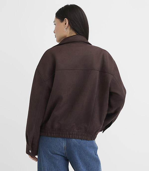 A woman in a brown jacket and blue jeans stands against a plain white background.