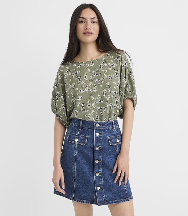A woman in a green paisley-patterned top and a denim skirt stands against a plain white background.