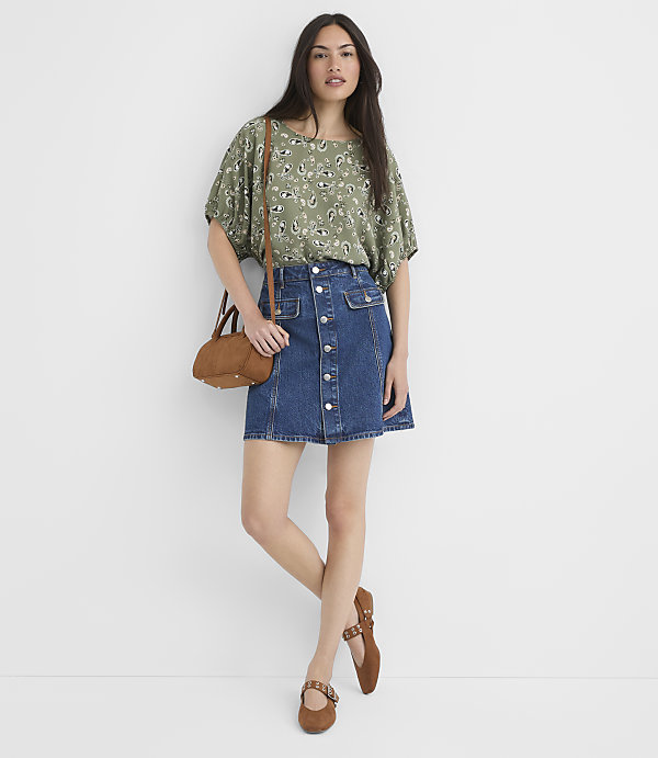 A woman in a floral top and denim skirt stands against a plain white background, with a brown crossbody bag and matching shoes.