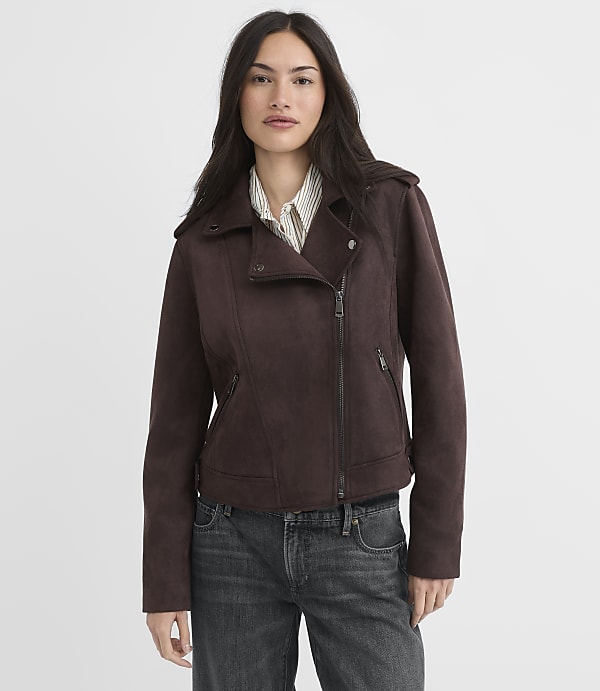 A woman in a brown suede moto jacket and jeans stands against a plain, light brown background.